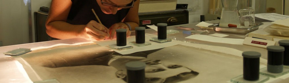 Image of Lisa Duncan performing art conservation treatments on an artwork showing a man's head and shoulders. On the artwork, several treatments are weighted under small stacks of blotter paper, plexiglass and weights.
